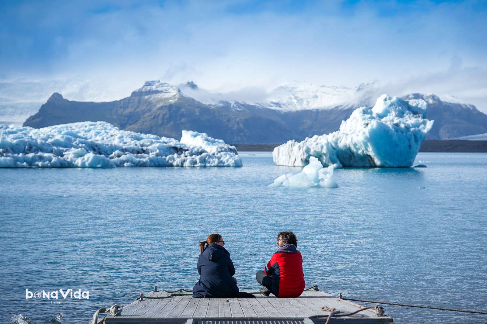 Dues persones assegudes a un moll mirant cap a un llac amb icebergs i muntanyes al fons.
