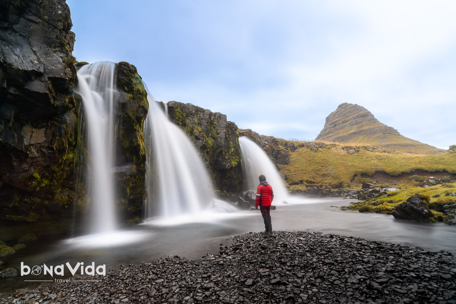 Una persona contemplant una cascada d'aigua en un paisatge natural, amb una muntanya al fons.