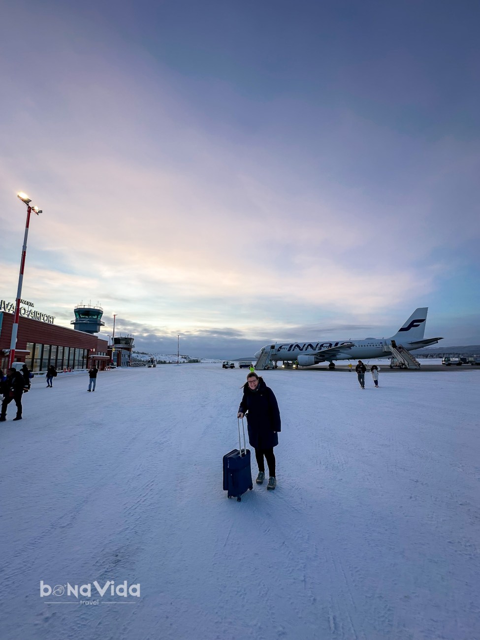 Una persona amb una maleta de rodets caminant per una pista de gel a prop d'un avió de Finnair i un aeroport, amb un cel de colors durant l'hora daurada.