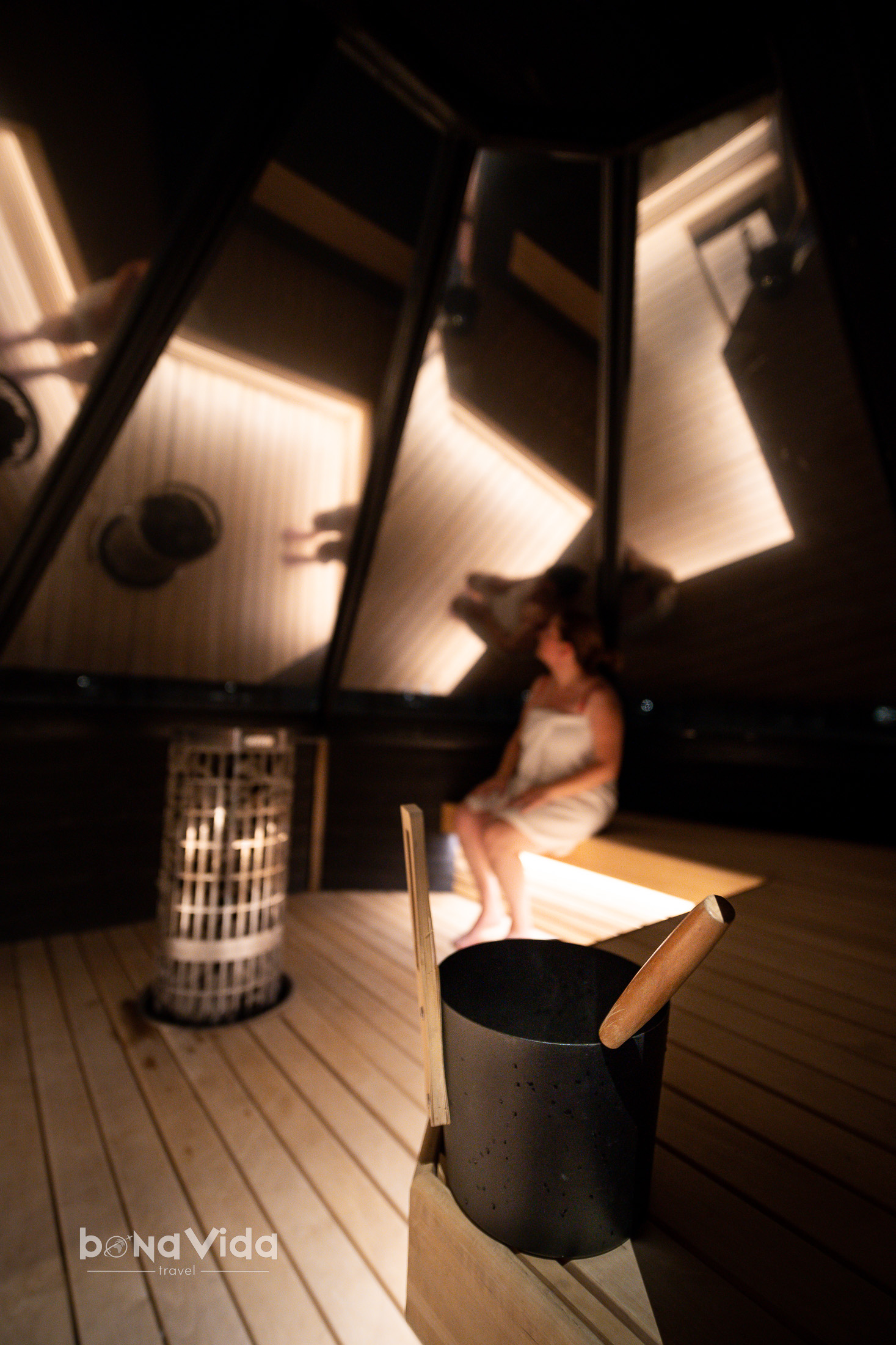 Interior d'una sauna amb una persona asseguda, amb un cub de pedra i un tassó de sauna a primer pla.