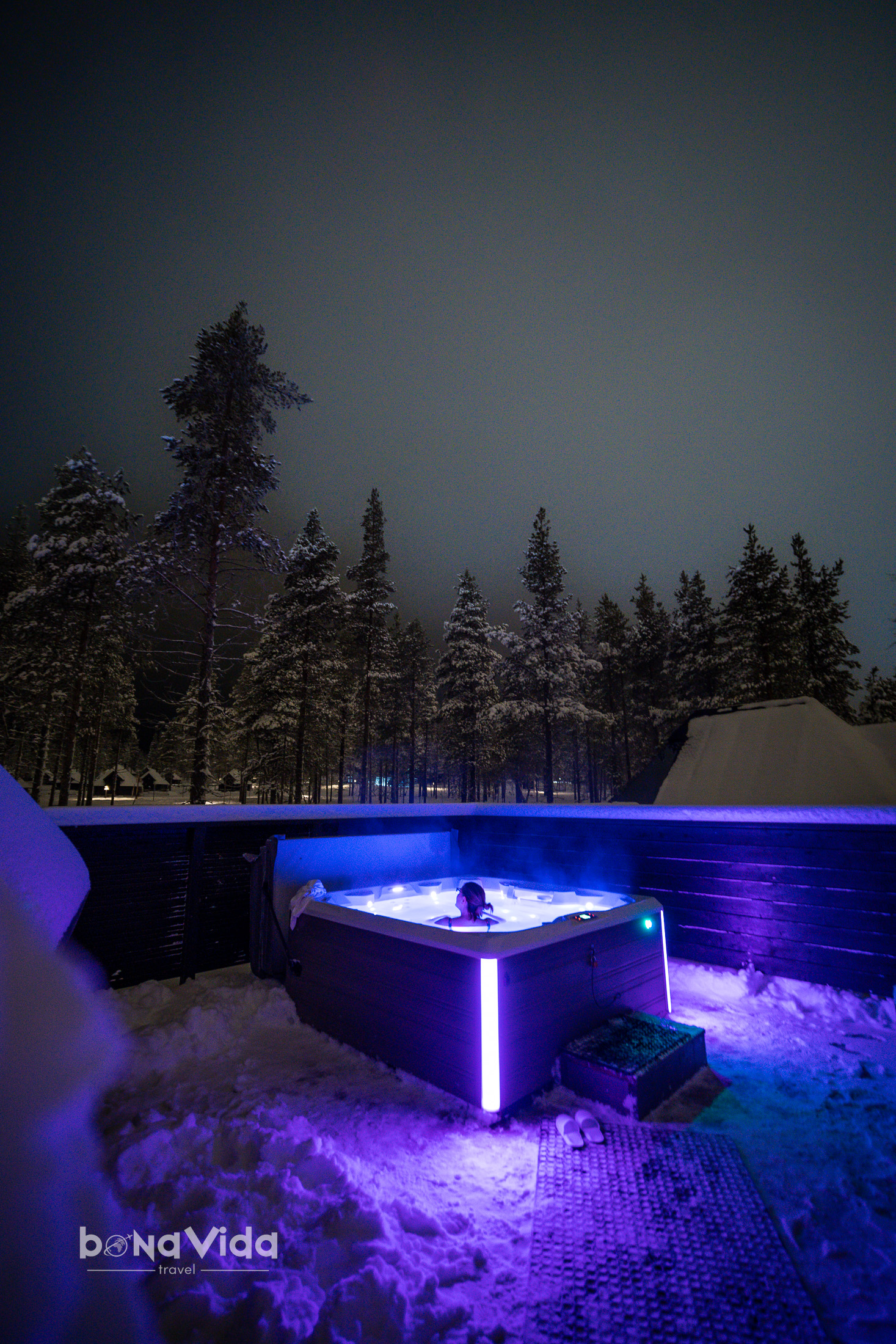 Una persona relaxant-se en una jacuzzi exterior envoltat de neu i arbres, amb il·luminació nocturna.