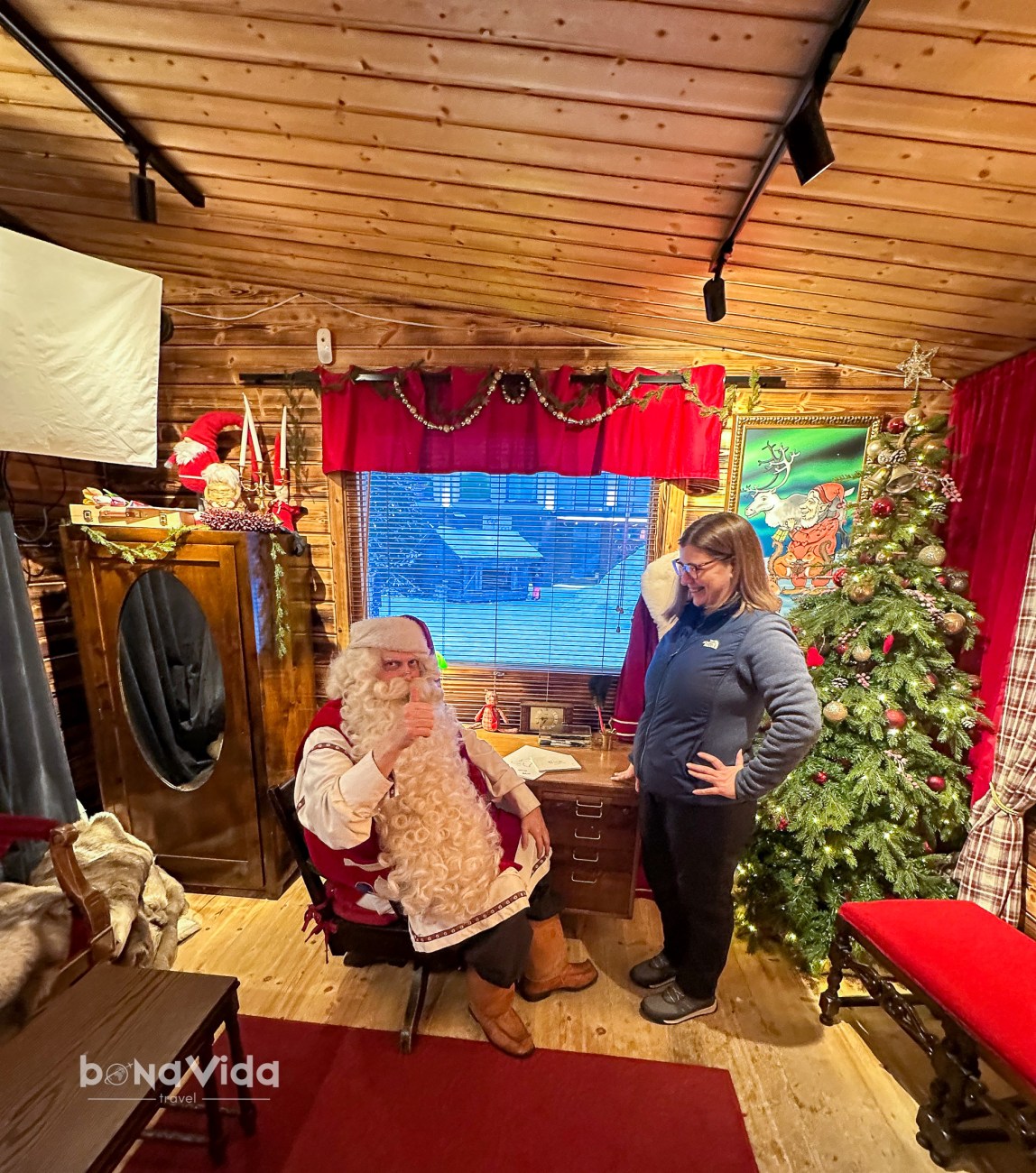Imatge d'un home vestit de Pare Noël assegut en una cadira, fent un gest de 'molt bé', mentre una dona amb ulleres somriu davant d'ell. La decoració inclou un arbre de Nadal i elements festius en una cabana de fusta.