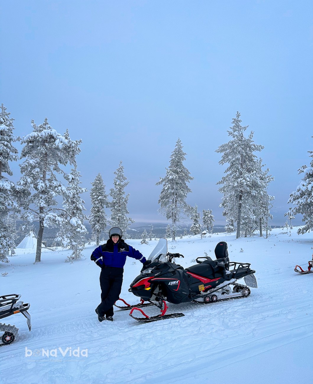 Una persona amb vestit d'esquí de peu al costat d'una moto de neu en un paisatge nevi amb arbres coberts de neu.
