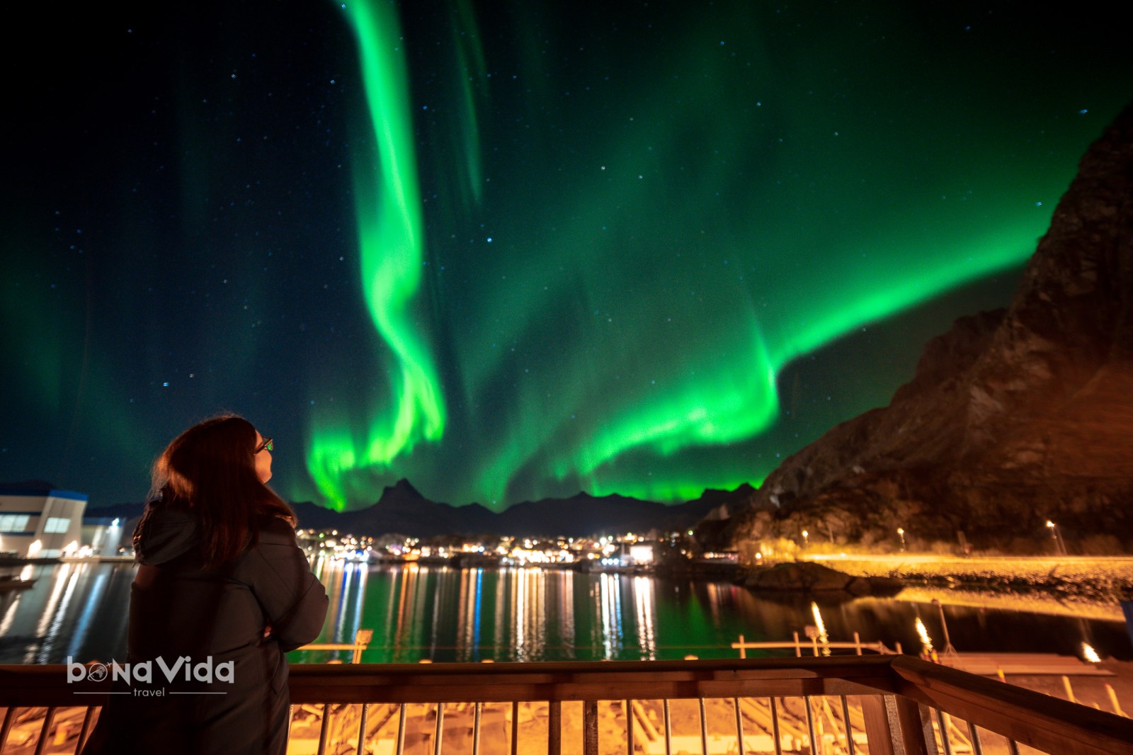 Una persona admirant les aurores boreals brillants sobre un paisatge nocturn, amb estrelles i un reflex d'il.luminació a la superfície d'un llac.