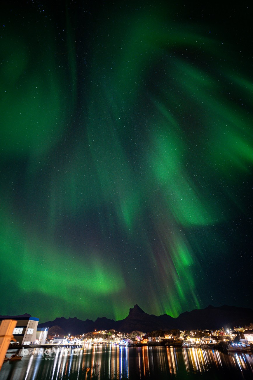 Aurora boreal brillant sobre un paisatge nocturn, amb muntanyes i un poble reflectit en l'aigua.