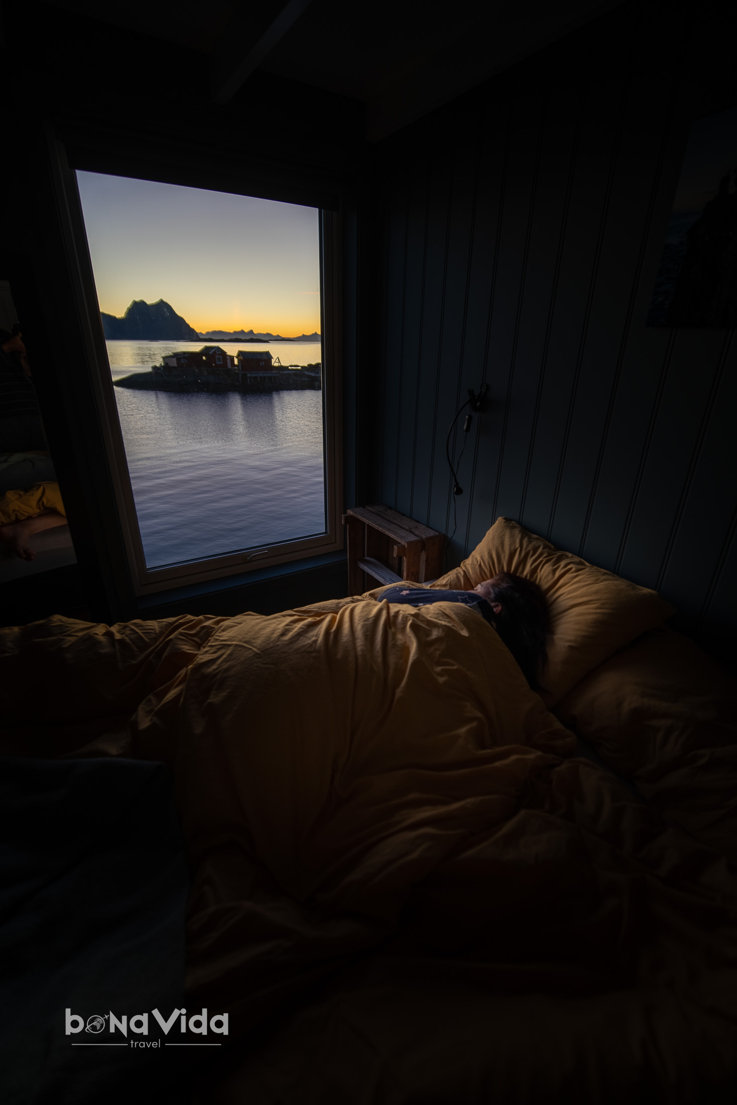 Una persona dormint en un llit amb roba de llit groga, amb una finestra que mostra una vista d'un paisatge tranquil amb una casa flotant sobre l'aigua al capvespre.