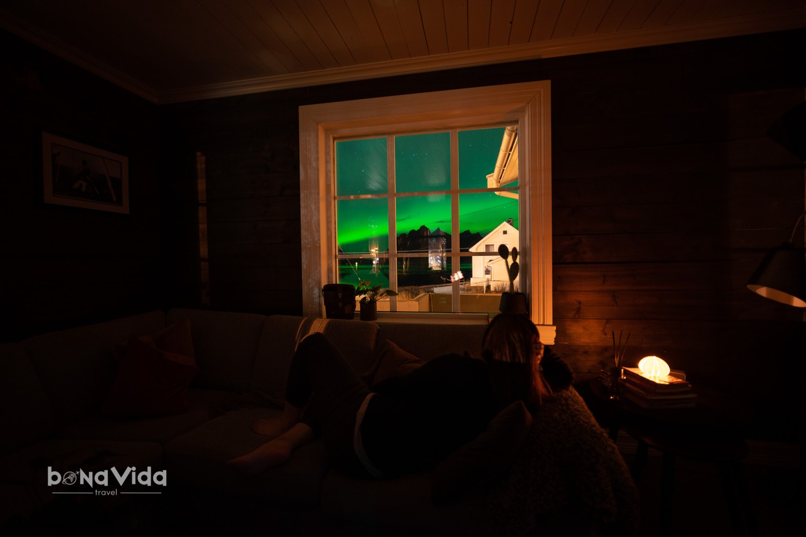 Una persona asseguda en un sofà, mirant per una finestra il·luminada amb una aurora boreal de fons i paisatge nocturn.