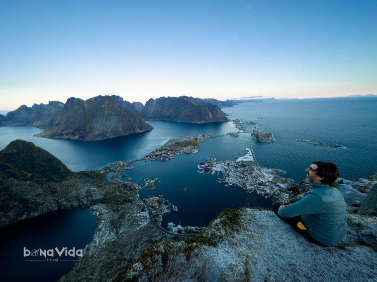 Un home se seu a la vora d'un penya-segat, contemplant una vista panoràmica de fiords i un petit poble costaner al fons, amb l'oceà en calma reflectint el cel.