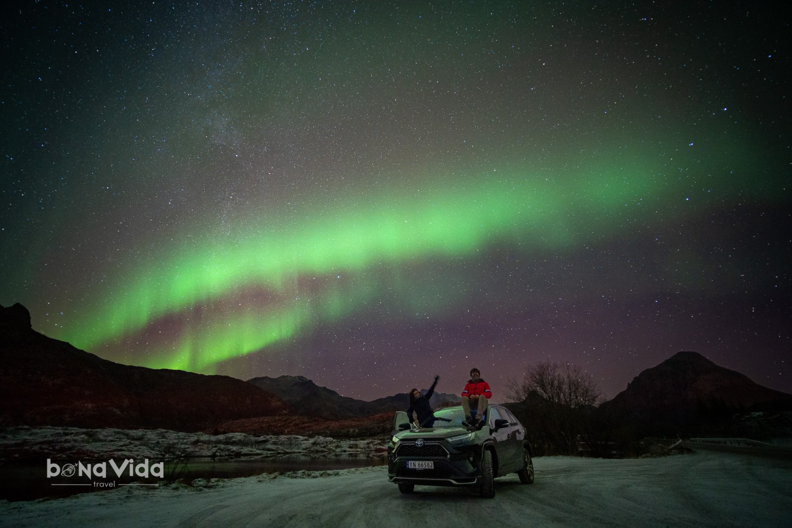 Dos persones assegudes sobre un cotxe sota l'aurora boreal amb estrelles de fons.