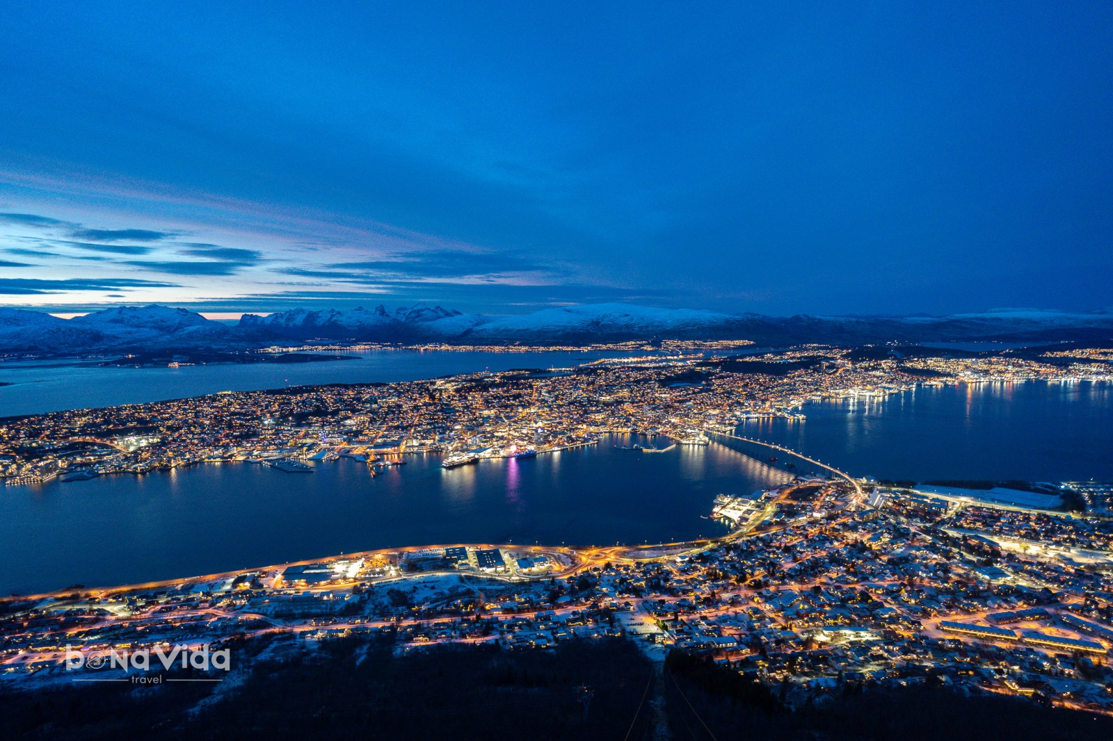 Vista aèria d'una ciutat il·luminada al vespre amb muntanyes nevades de fons i un llac reflexant les llums.