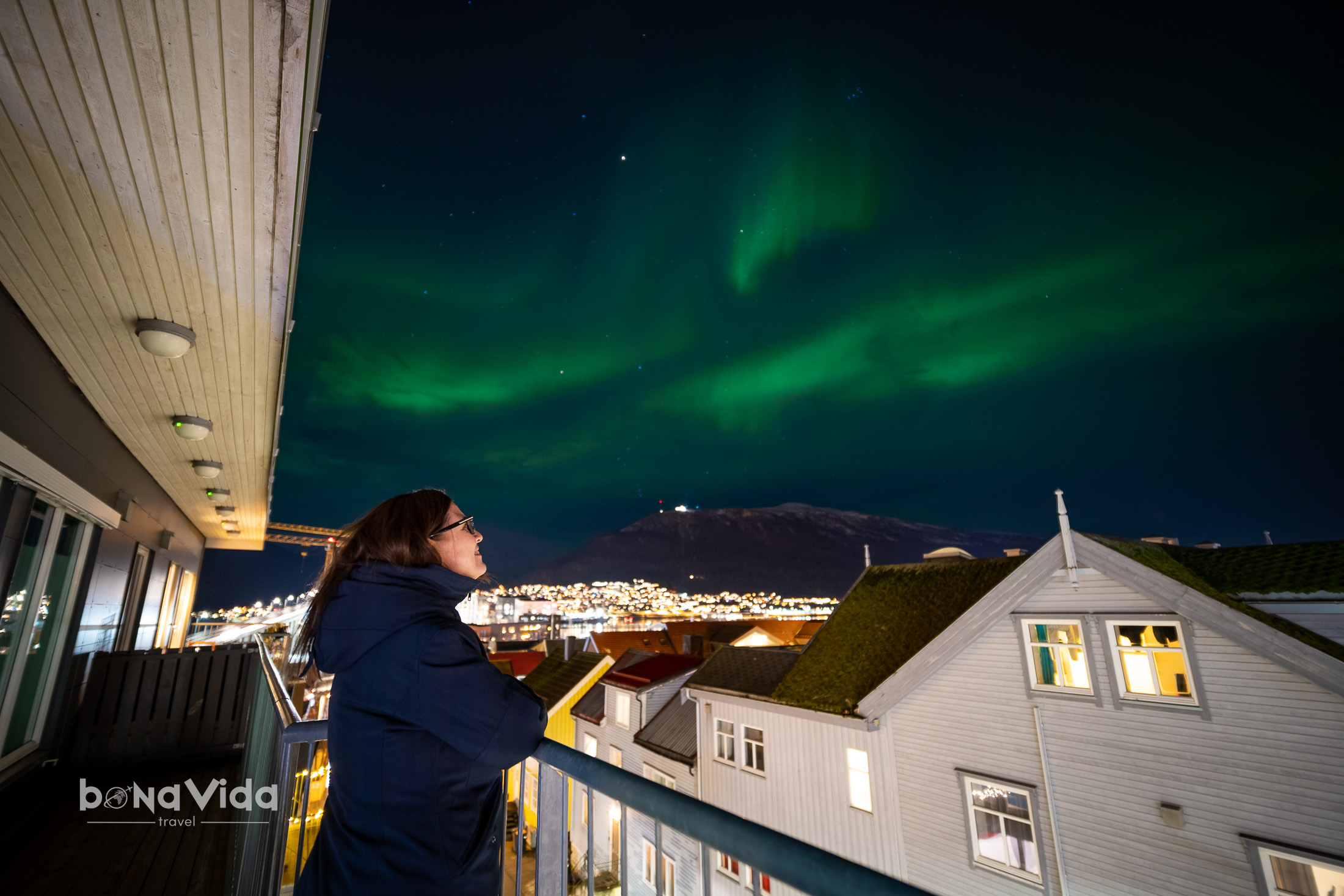 Una dona mirant l'aurora boreal des d'un balcó durant la nit, amb ciutats il·luminades al fons.