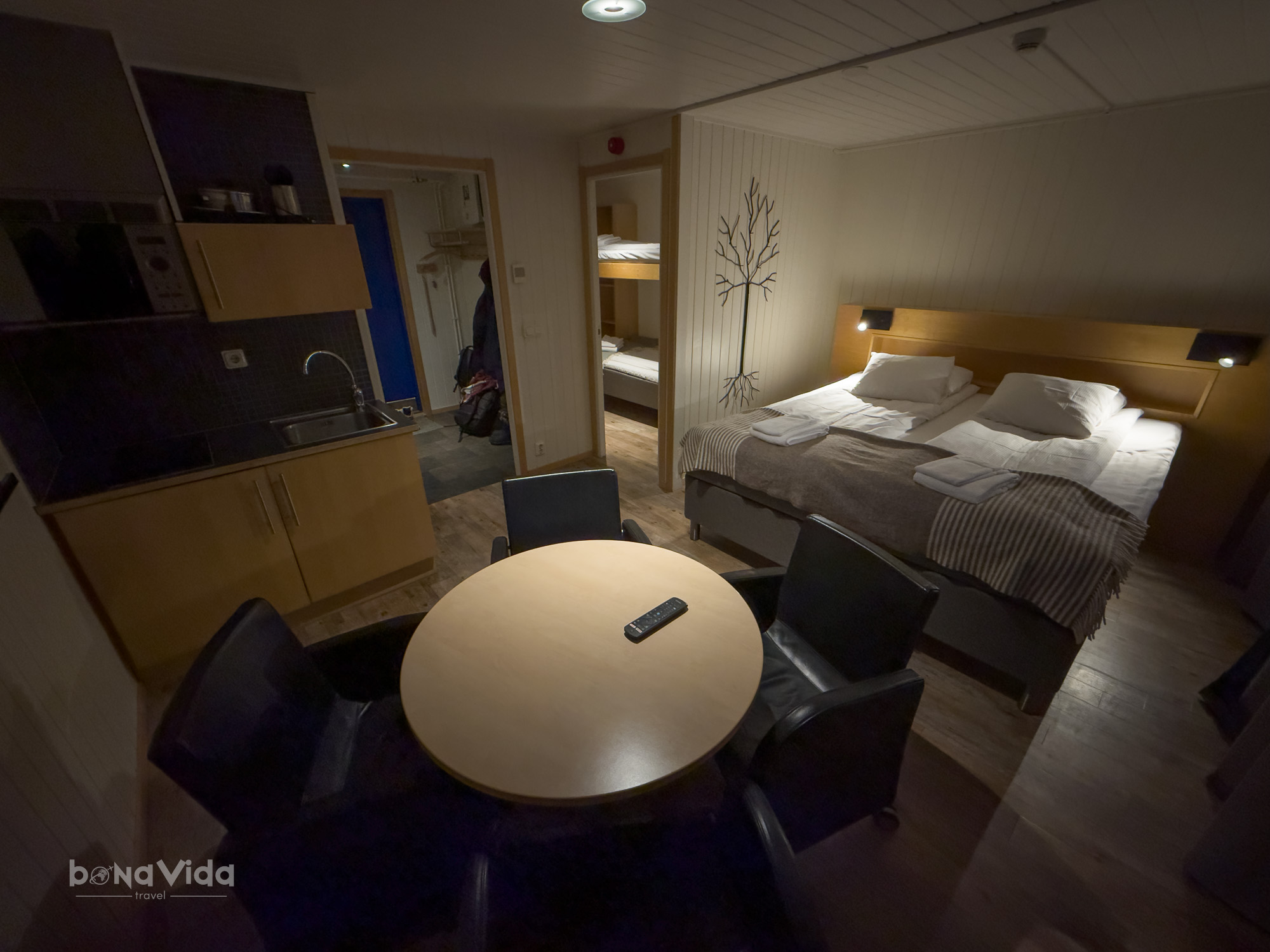 Interior d'un apartament amb cuina, una taula rodona i cadires, i un llit doble amb llençols blancs. S'hi poden veure llits superposats a la part posterior.